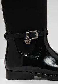 Botte de pluie noire brillante avec bride à la cheville, boucle argentée et pendentif rond en argent avec les initiales "MK".