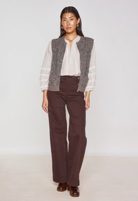 Gilet in maglia grigia con texture sopra una blusa bianca a righe, abbinato a pantaloni marroni a vita alta e scarpe marroni.