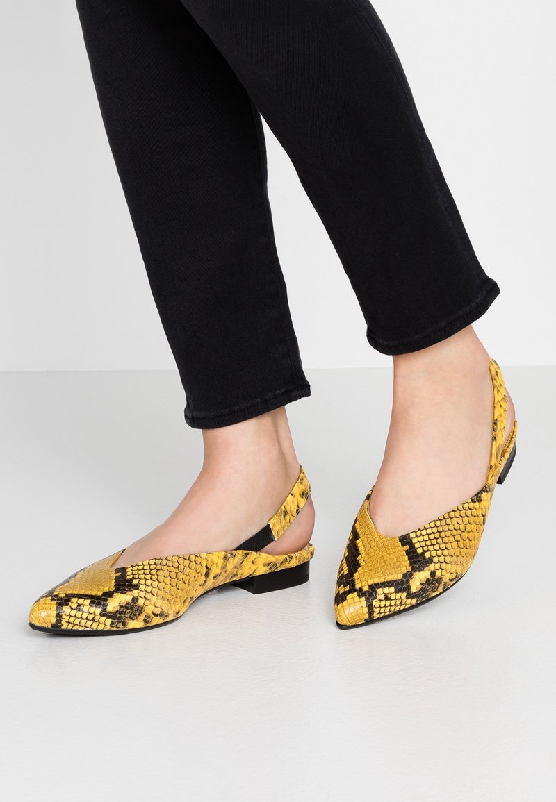 Chaussures plates slingback jaune et noir à motif peau de serpent, portées avec un pantalon court noir sur un sol clair et un fond uni.