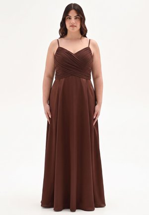 Donna in piedi con un vestito marrone lungo fino al pavimento, con spalline sottili e corpetto plissettato, su uno sfondo bianco semplice.