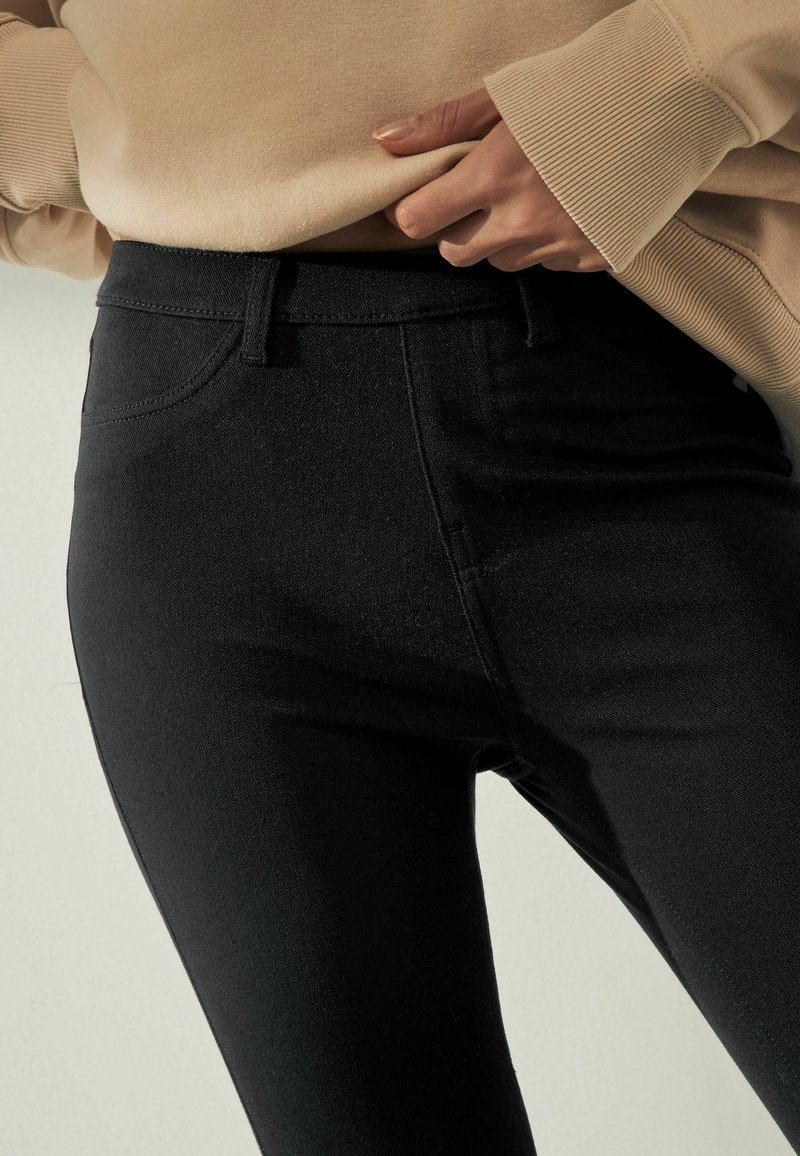 Zwarte skinny jeans met een gladde textuur, voorzien van vijf zakken, een klassieke tailleband en minimale stikseldetails, gedragen met een beige top.