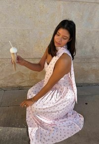 Femme en robe florale rose à genoux sur une surface en pierre tenant un cornet de glace fondant avec une cuillère devant un mur beige.