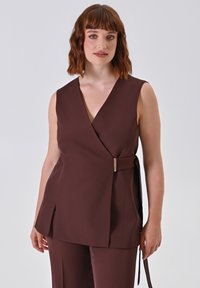 Donna che indossa un top avvolgente senza maniche di colore marrone scuro con dettaglio della fibbia laterale e pantaloni coordinati, in piedi di fronte a uno sfondo chiaro e semplice.