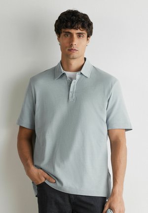 Homme aux cheveux bouclés et noirs portant un polo gris clair à manches courtes par-dessus un maillot blanc, debout contre un mur uni.