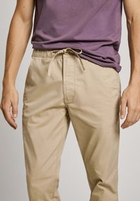 Pantalones informales de color beige claro con cintura elástica y cordón. La tela es suave con detalles de costura sutiles.