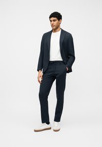 Άνδρας που στέκεται μπροστά σε λευκό φόντο, φορώντας μπλε κοστούμι, λευκό t-shirt και λευκά αθλητικά παπούτσια με καφέ σόλες.