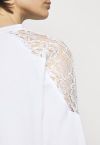 Vit sweatshirt med spetsdetaljer på axlarna, med en texturerad design. Spetsen har ett blommönster och är semi-transparent.