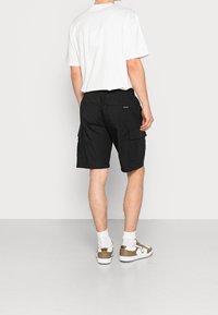 Černé cargo šortky s více kapsami, elastickým pasem a délku ke kolenům, spárované s bílým tričkem a teniskami s hnědými akcenty.