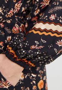 Tissu noir à motif floral, avec des rayures décoratives orange et beige, présentant des poignets élastiques et une poche pour un détail supplémentaire.