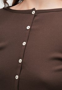 Haut en tissu côtelé marron avec un col rond et six boutons blancs sur le devant. Présente une finition texturée et douce.