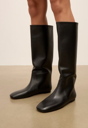 Jambes portant de hautes bottes en cuir noir lisse avec une semelle plate, debout sur un sol beige devant un fond uni.