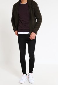 Dunkelgrüne gestrickte Strickjacke über einem burgunderfarbenen Pullover, weißen T-Shirt, schwarzen Skinny-Jeans und weißen Sneakers mit rosa Akzenten.