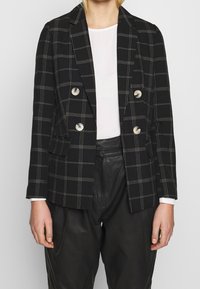 Två-knapprad svart kavaj med vit rutmönster, med silverbiseringar och spetsiga slag. Matchad med en vit topp och läderbyxor.