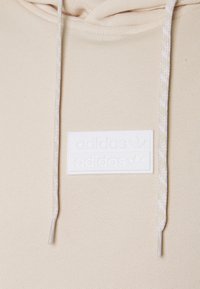 Beige huvihiiri, jossa on teksturoitu kangas, valkoinen Adidas-logo ja vaaleat, hienovaraisella kuviolla varustetut kiristysnauhat.