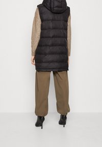 Svart pufferväst med huva, med en quiltad design, kombinerad med en beige tröja och olivfärgade vida byxor. Svarta klackstövlar fulländar looken.