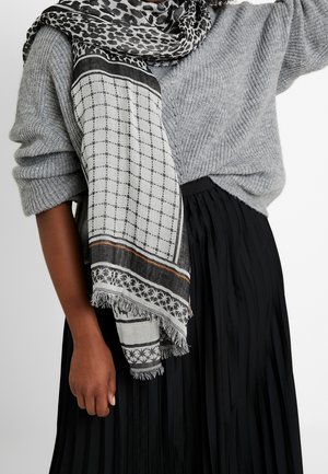 Femme portant un pull en maille gris, une jupe plissée noire et une écharpe à motifs noir et blanc drapée sur l'épaule et autour du cou.