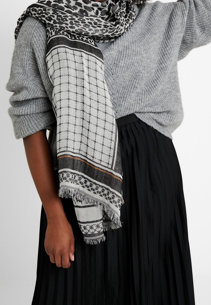 Femme portant un pull en maille gris, une jupe plissée noire et une écharpe à motifs noir et blanc drapée sur l'épaule et autour du cou.
