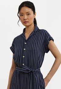 Vestido de riscas azul-marinho com mangas curtas enroladas, frente com botões, bolso no peito e cinto amarrado na cintura. Textura de tecido suave.