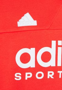 Felpa rossa con logo bianco "adidas" e testo "SPORT". Presenta un piccolo logo trefoil bianco in alto a sinistra. Tessuto morbido e texture.