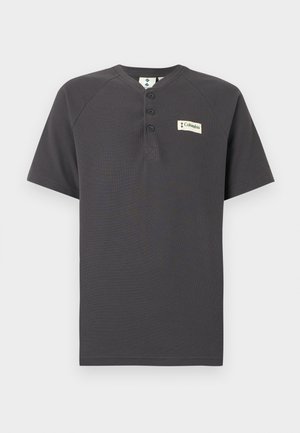 Camisa Henley de manga corta color gris oscuro con tres botones y un pequeño parche con el logo de Columbia en el pecho izquierdo sobre un fondo blanco.