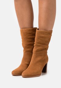 Botas de ante marrón suave hasta la mitad de la pantorrilla con tacones cuadrados, usadas sobre piernas desnudas, con un sutil detalle de costura en los lados exteriores.