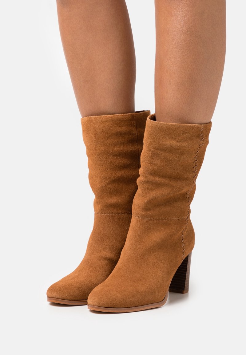 Botas de ante marrón suave hasta la mitad de la pantorrilla con tacones cuadrados, usadas sobre piernas desnudas, con un sutil detalle de costura en los lados exteriores.