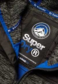 Sort og gråt tekstureret stof med lyseblå accenter; indeholder en fremtrædende sort mærkat med "Super Dry," størrelse "L" og bjerglogo.