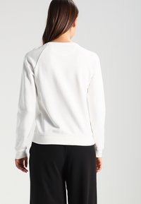 Mujer con cabello largo castaño, vestida con una camiseta blanca de manga larga lisa y pantalones negros holgados, de pie frente a un fondo gris claro liso.