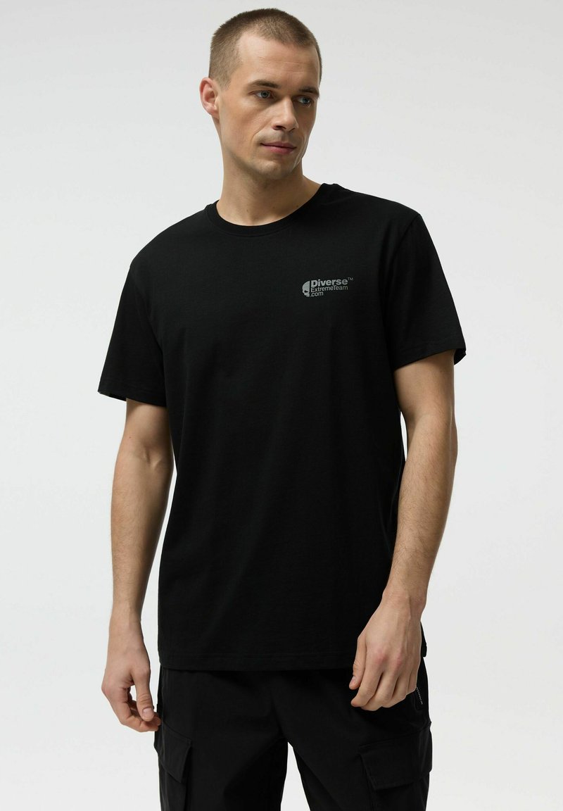 Mężczyzna ubrany w czarną koszulkę z okrągłym dekoltem z logo "Diverse ExtremeTeam" na klatce piersiowej oraz czarne krótkie spodnie typu cargo, stojący na jednolitym tle.