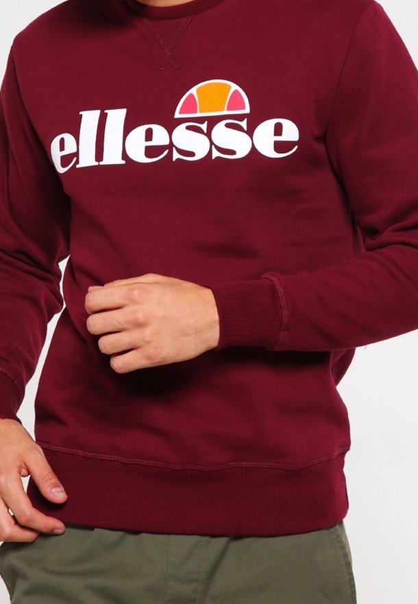 Sweat-shirt bordeaux avec logo blanc "ellesse" et demi-cercle orange au-dessus du texte, porté par une personne ajustant sa manche.
