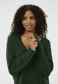Cardigan verde tricotat cu decolteu în V, manșete cu nervuri și nasturi în față. Textură moale, croială lejeră, cu un design relaxat.