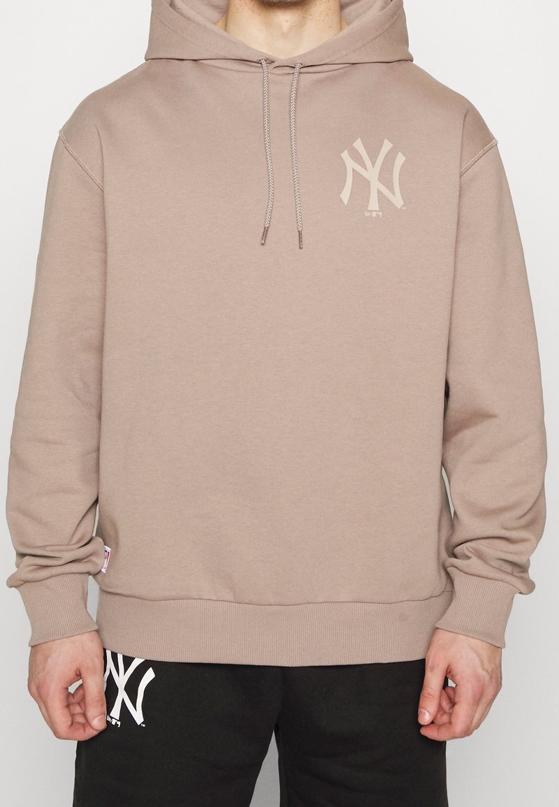 Personne portant un sweat à capuche beige avec un logo NY clair sur la poitrine et un pantalon noir avec un logo NY blanc près de la poche.