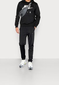 Sudadera negra con el logo blanco de Nike, pantalones negros a juego, bolso cruzado gris y zapatillas blancas con detalles en gris y amarillo.
