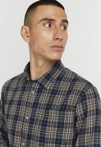Braun und marineblau karierter Button-Down-Hemd mit Kragen, ausgestattet mit einer Brusttasche und aus leichtem Stoff, der ein karierter Muster zeigt.