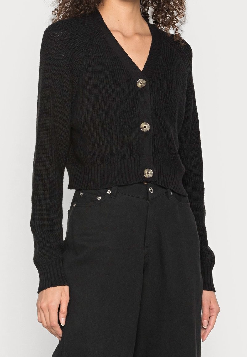 Femme portant un cardigan côtelé noir à gros boutons associé à un pantalon noir taille haute, sur un fond uni.