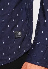 Marineblauwe shirt met kleine witte geometrische patronen, met een knoopsluiting kraag en een zichtbaar zwart label aan de onderkant links. Van katoen.