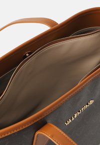 Didelis juodas moteriškas krepšys su rudos odos akcentais, auksiniu logotipu ir užtrauktuku. Lygi tekstūra, erdvi vidaus dalis.