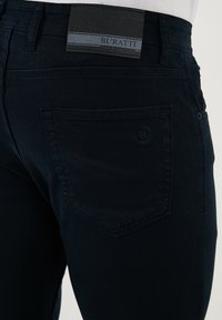 Temno modri jeans iz denima imajo zadnji žep, reliefno logotipno podrobnost in črno usnjeno oznako s sivo črtasto dekoro na pasu.