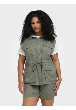 Kvinde med krøllet hår iført en grøn vest med lommer og matchende shorts over en hvid kortærmet skjorte, stående mod en lys baggrund.