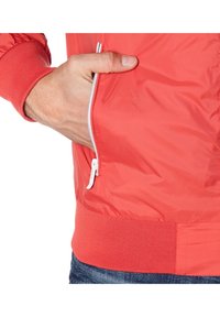 Veste zippée rouge avec une finition texturée, dotée d'une fermeture éclair blanche et de poignets en tricot côtelé. Main partiellement visible dans la poche latérale.