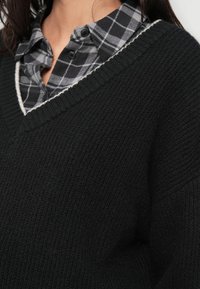 Melns adīts džemperis ar V veida izgriezumu, valkāts virs pelēkas un melnas pārbaudītas flaneļa krekla ar pogām un teksturētu audumu.