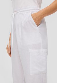 Weiße Leinenhose mit elastischem Bund, Kordelzug, Seitentaschen und einer aufgesetzten Tasche am Bein. Leichte und atmungsaktive Textur.