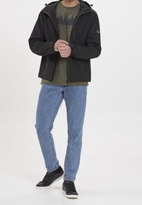 Mężczyzna w czarnej kurtce z kapturem, oliwkowej koszulce z grafiką, jasnoniebieskich dżinsach i czarnych sneakersach na białym tle.