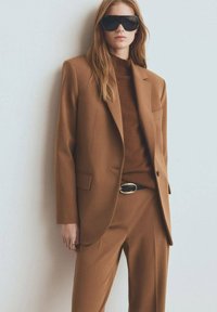 Traje marrón a medida con blazer de un solo botón y pantalones a juego, cuello alto marrón acanalado debajo y gafas de sol negras oversized.