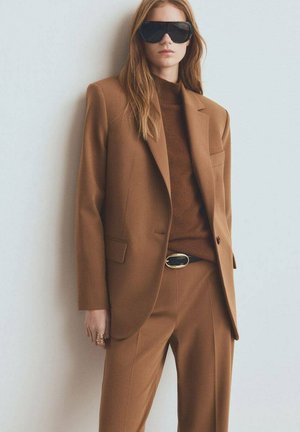 Traje marrón a medida con blazer de un solo botón y pantalones a juego, cuello alto marrón acanalado debajo y gafas de sol negras oversized.