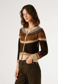 Cardigan en tricot marron et crème avec un motif en zigzag, fermeture boutonnée et rayures contrastées. Manches longues, finitions côtelées aux poignets et à l'ourlet.