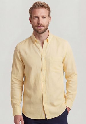 Man met lichte baard die een lichtgeel overhemd met knopen en lange mouwen draagt en donkere broek, staand met één hand in de zak.