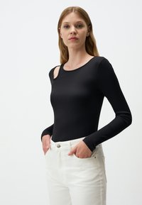 Top lungo a maniche ribbed nere con dettagli cut-out sulle spalle, abbinato a pantaloni chiari con chiusura a bottone.