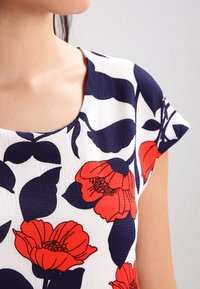 Top corto de flores con mangas cortas y escote redondo. Base blanca con grandes flores rojas y azules marinos y follaje. Tejido texturizado con un patrón sutil.