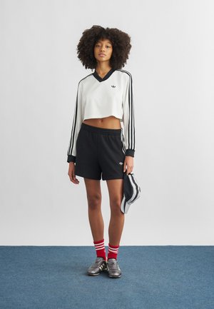 Młoda kobieta o kręconych włosach ubrana w czarno-białe sportowe ubrania marki Adidas, czerwone skarpetki w paski, metaliczne sneakersy, trzymająca dopasowaną czapkę.